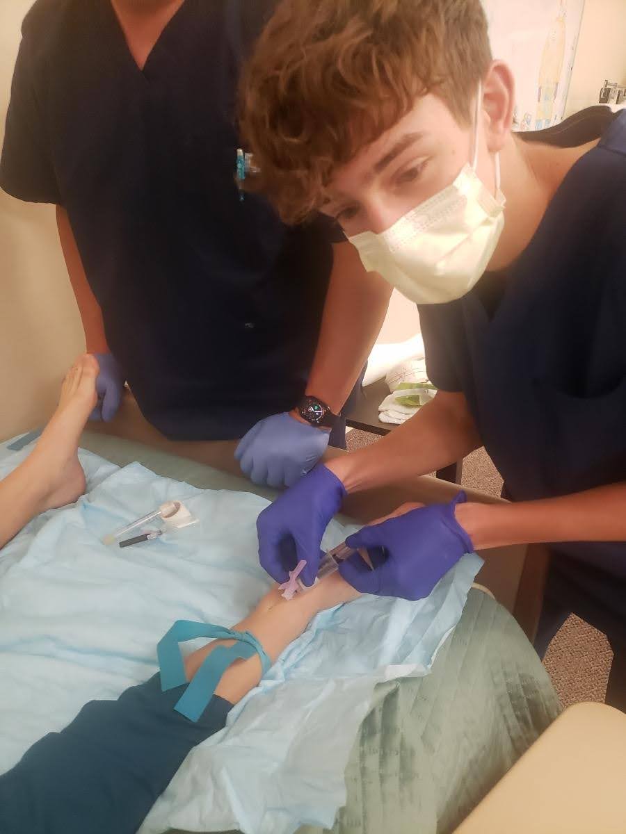 Medical students in scrubs practice IV insertion on a model's arm, demonstrating clinical techniques in a training environment.