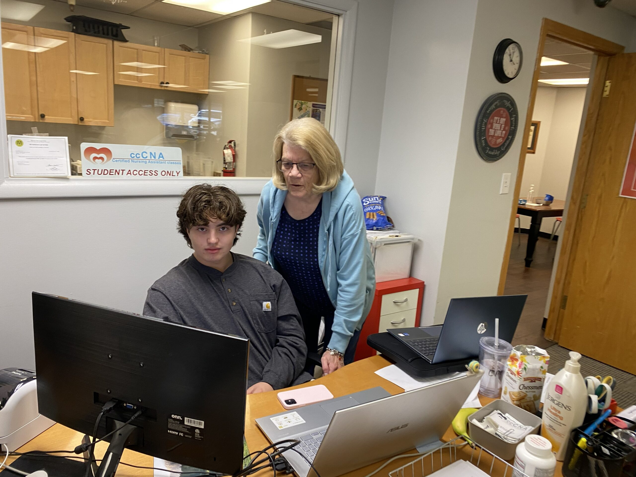 A woman assists a student at a desk with multiple computers and supplies in a classroom setting, emphasizing a supportive learning environment.