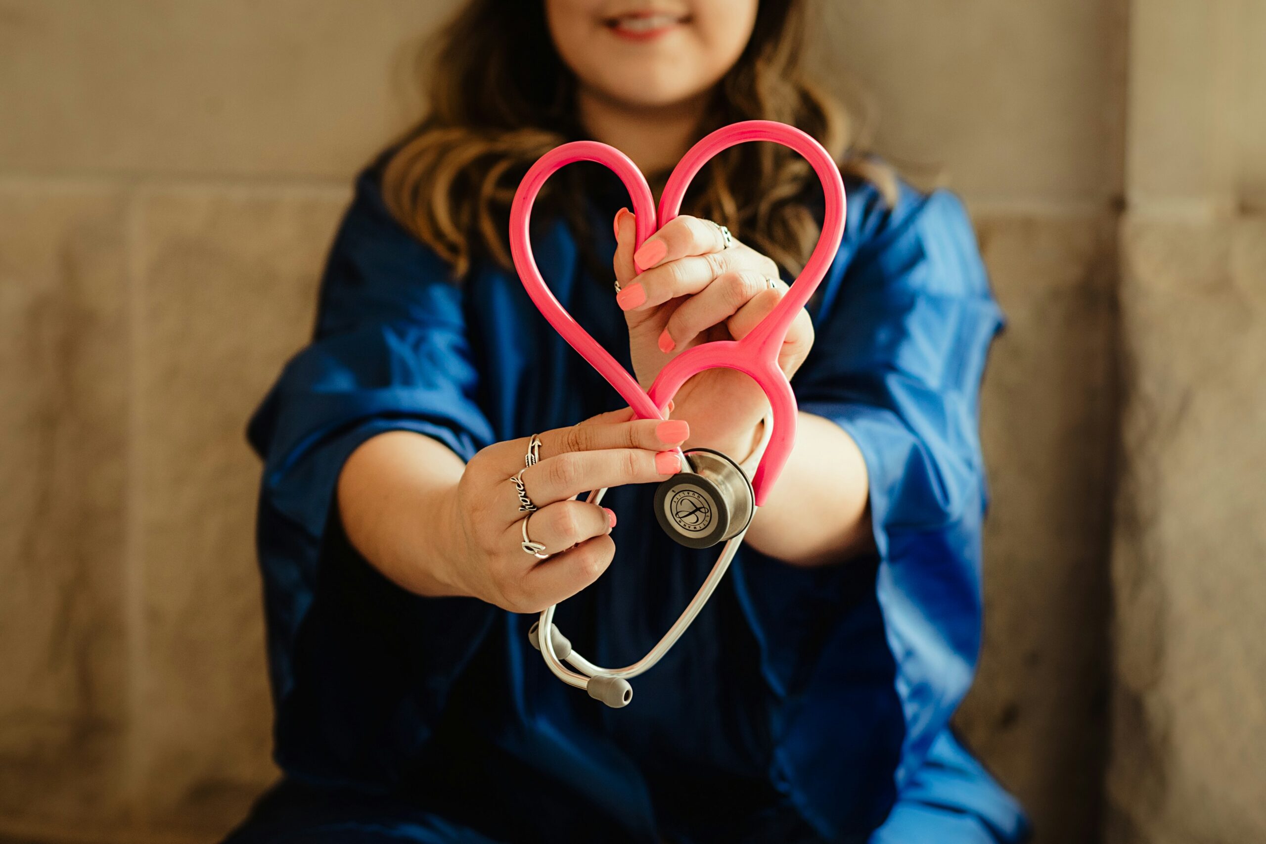 A person in a blue robe holds a pink stethoscope shaped like a heart, symbolizing caring in healthcare. The image emphasizes compassion in medicine.