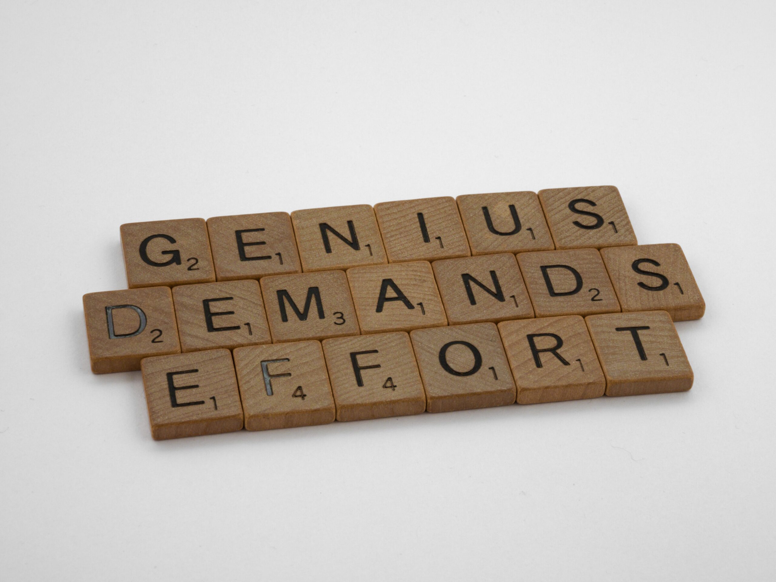Wooden letter tiles spell out "GENIUS DEMANDS EFFORT" against a plain background, emphasizing the message about perseverance and dedication.